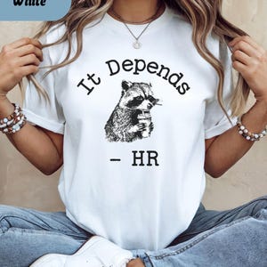 May include: White t-shirt with the text "It Depends" above a black and white illustration of a raccoon holding a cup. Below the raccoon is the text "- HR". The shirt has short sleeves.