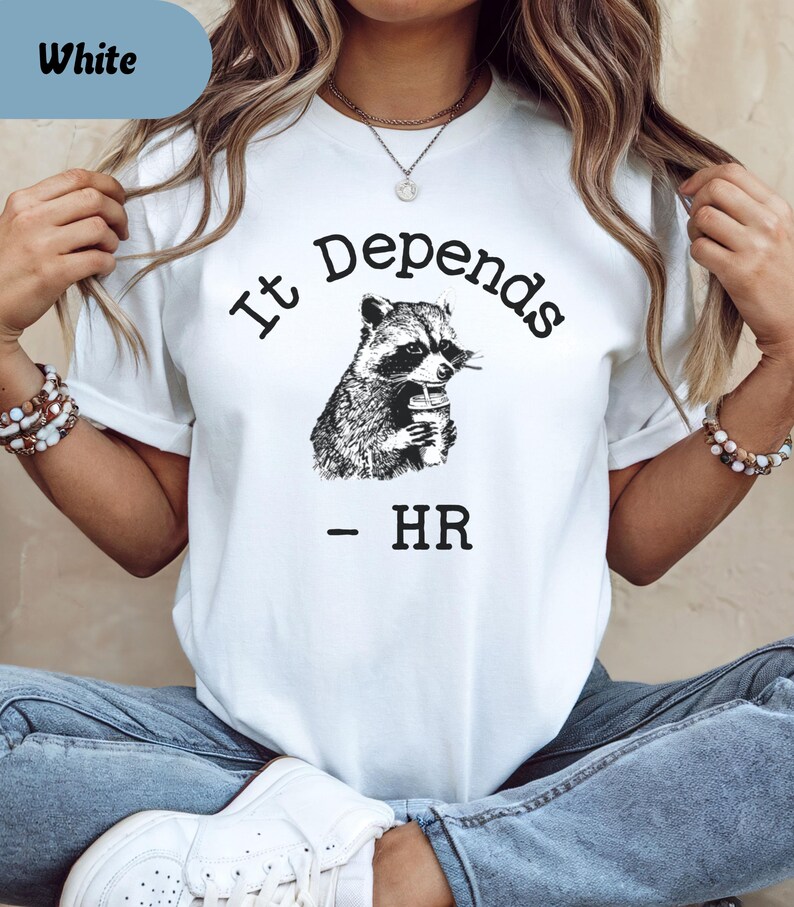 May include: White t-shirt with the text "It Depends" above a black and white illustration of a raccoon holding a cup. Below the raccoon is the text "- HR". The shirt has short sleeves.