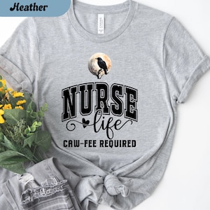May include: Athletic heather gray t-shirt with the words "NURSE life CAW-FEE REQUIRED" in black. A black raven is perched on a branch in front of a full moon. The words "Athletic Heather" are in a blue box.