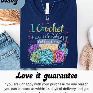 May include: Navy blue t-shirt with the text "I Crochet but my favorite hobby is collecting YARN" and colorful yarn balls. A silver necklace with a sun pendant is visible. The shirt is folded on top of blue jeans.