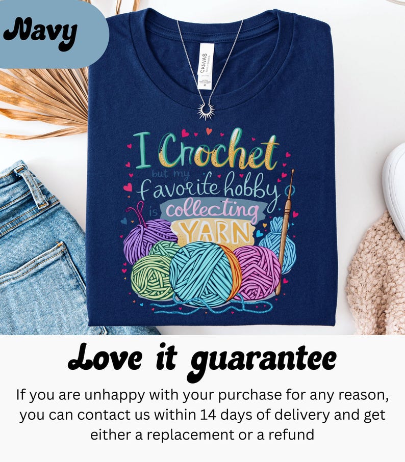 May include: Navy blue t-shirt with the text "I Crochet but my favorite hobby is collecting YARN" and colorful yarn balls. A silver necklace with a sun pendant is visible. The shirt is folded on top of blue jeans.