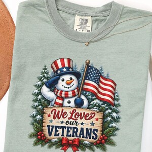 May include: A bay-coloured t-shirt featuring a graphic of a snowman wearing a patriotic top hat and scarf, saluting while holding an American flag. Below the snowman is a wooden sign with the text We Loves our VETERANS, decorated with pine.