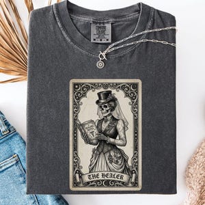 May include: Dark gray t-shirt with a tarot card graphic. The card features a skeleton in Victorian attire, holding a book labeled "Healing Notes." The card is framed with ornate scrollwork, and the banner reads "The Healer."