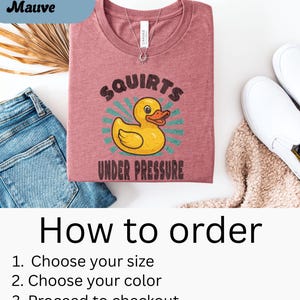 以下が含まれることがあります： 黄色いアヒルのイラストと「SQUIRTS UNDER PRESSURE」の文字が入った、ヘザーモーヴのTシャツ。Tシャツは、青いジーンズ、白い靴、ベージュのセーターと一緒に表示されています。