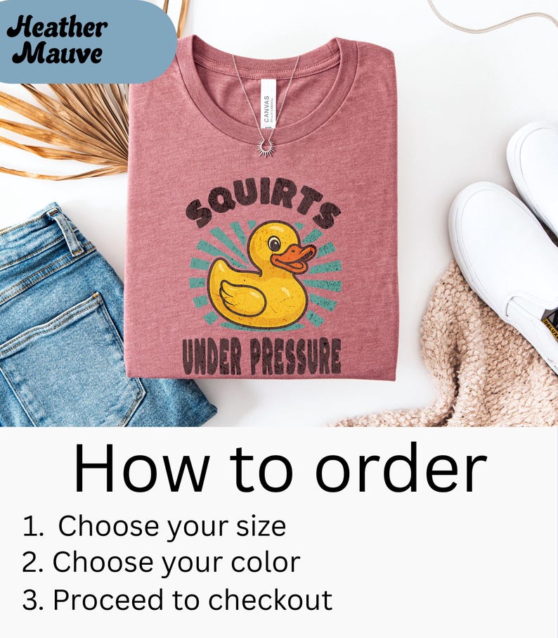 以下が含まれることがあります： 黄色いアヒルのイラストと「SQUIRTS UNDER PRESSURE」の文字が入った、ヘザーモーヴのTシャツ。Tシャツは、青いジーンズ、白い靴、ベージュのセーターと一緒に表示されています。