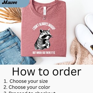 May include: A heather mauve t-shirt with the text "I DON'T ALWAYS WHOOP BUT WHEN I DO THERE IT IS" and a graphic of a raccoon holding a drink. The shirt is folded and displayed with blue jeans, white shoes, and a beige sweater.