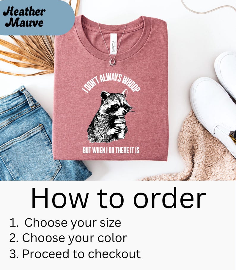 May include: A heather mauve t-shirt with the text "I DON'T ALWAYS WHOOP BUT WHEN I DO THERE IT IS" and a graphic of a raccoon holding a drink. The shirt is folded and displayed with blue jeans, white shoes, and a beige sweater.
