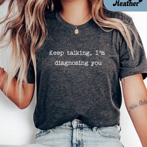May include: Dark grey heather t-shirt with the text "Keep talking, I'm diagnosing you" in white. The shirt is a dark grey colour and has a crew neck. The shirt is made of a soft material.
