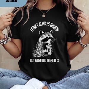 May include: Black t-shirt featuring a raccoon graphic holding a beverage. The text "I DON'T ALWAYS WHOOP BUT WHEN I DO THERE IT IS" is printed above and below the raccoon illustration. The shirt is a crew neck.