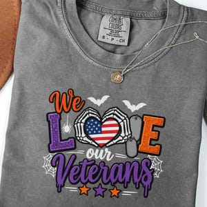 May include: A charcoal grey t-shirt featuring a graphic with the text We Love our Veterans. The design includes skeleton hands forming a heart shape around an American flag, two silver dog tags, a spider, bats, and spiderwebs in orange and purple.