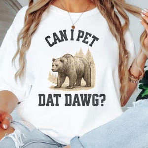 May include: White t-shirt with the text "CAN I PET DAT DAWG?" above and below a sepia-toned illustration of a bear. The bear is standing in front of a forest scene. The shirt is a casual, unisex style.