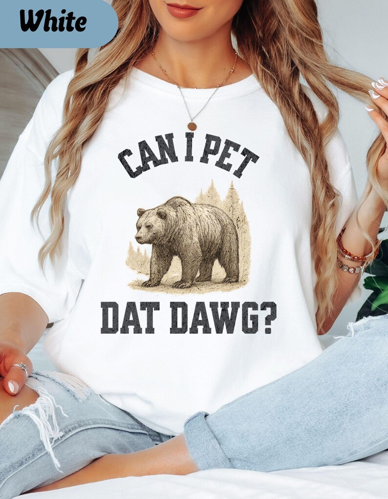 May include: White t-shirt with the text "CAN I PET DAT DAWG?" above and below a sepia-toned illustration of a bear. The bear is standing in front of a forest scene. The shirt is a casual, unisex style.
