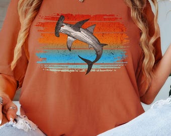 May include: A rust-colored t-shirt with a vintage-style graphic of a hammerhead shark swimming in front of a sunset. The graphic has a distressed look with faded colors.