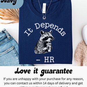 May include: Navy blue t-shirt with the text "It Depends" above a raccoon illustration holding a beverage. Below the raccoon is the text "- HR". A silver necklace with a sun pendant is visible.