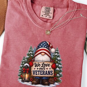 May include: A crimson t-shirt featuring a graphic of a gnome wearing a stars and stripes hat, holding a sign that reads We Love our VETERANS. The gnome stands in snow with pine trees, a lantern, and holly berries. The shirt has a Comfort Colors label.