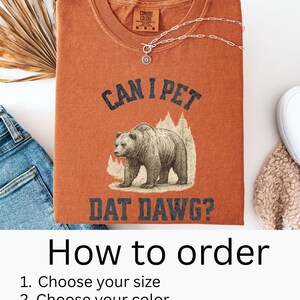 May include: A burnt orange t-shirt with the text "CAN I PET DAT DAWG?" and a graphic of a bear. The shirt also has the Comfort Colors logo and a silver necklace. The image includes instructions on how to order.