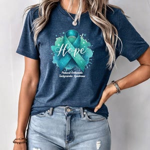 May include: A navy blue t-shirt with a teal and blue ribbon and the word "Hope" in a white font. The ribbon is surrounded by a watercolor-style blue and green splatter. The text "Postural Orthostatic Tachycardia Syndrome" is printed below the ribbon.