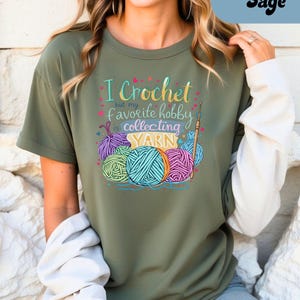 May include: Sage green t-shirt with the text "I Crochet but my favorite hobby is collecting YARN." The graphic features colorful yarn balls and a crochet hook. The shirt has short sleeves and a relaxed fit.