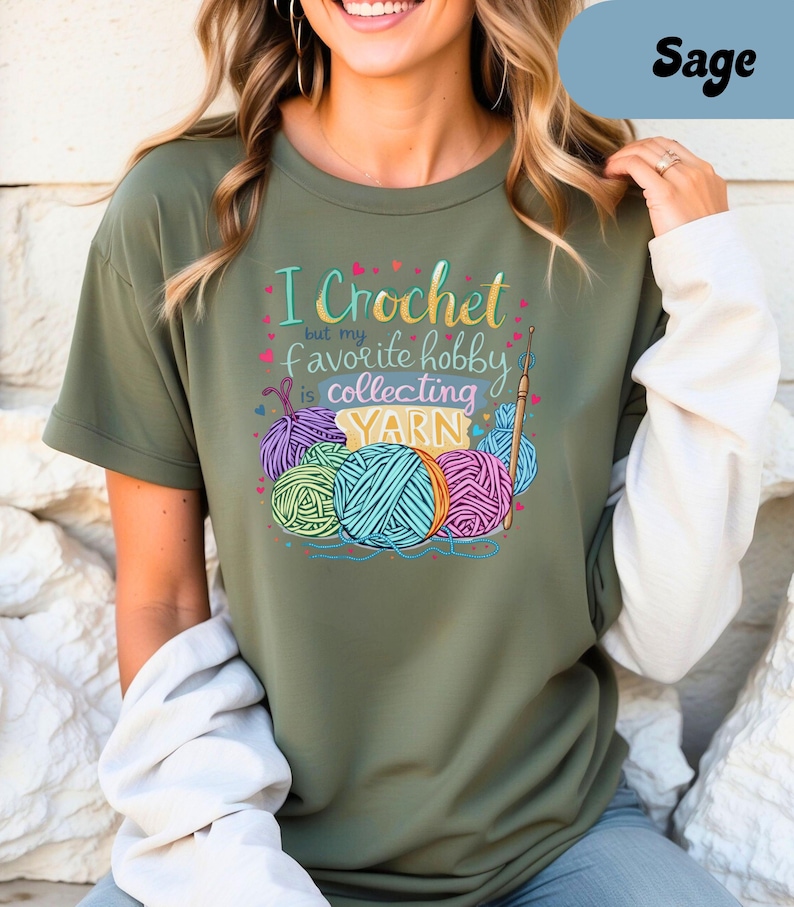 May include: Sage green t-shirt with the text "I Crochet but my favorite hobby is collecting YARN." The graphic features colorful yarn balls and a crochet hook. The shirt has short sleeves and a relaxed fit.