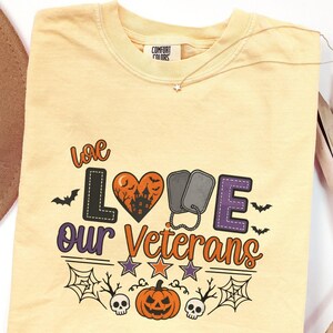 May include: A butter yellow t-shirt featuring a graphic with the text "we LOVE our Veterans". The design includes a heart-shaped haunted house, two military dog tags, bats, stars, spider webs, skulls, and a jack-o'-lantern in orange, purple, and black.