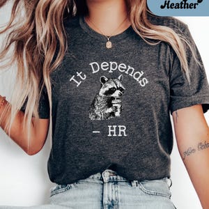 May include: Dark grey heather t-shirt with the text "It Depends" above a black and white illustration of a raccoon holding a cup. Below the raccoon is the text "- HR".