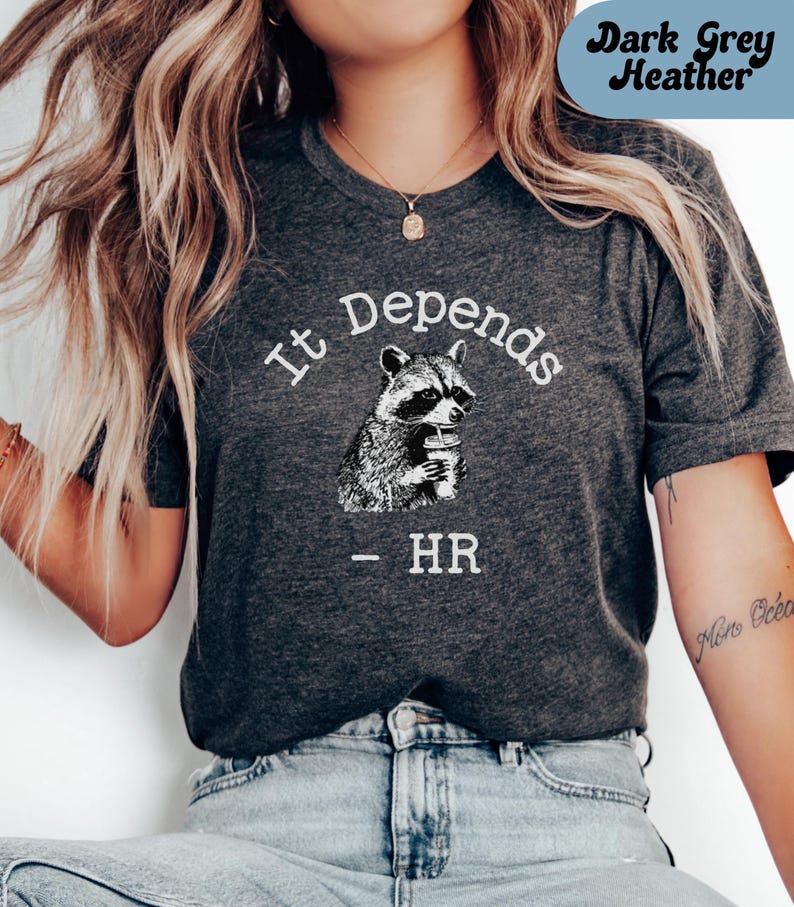 May include: Dark grey heather t-shirt with the text "It Depends" above a black and white illustration of a raccoon holding a cup. Below the raccoon is the text "- HR".