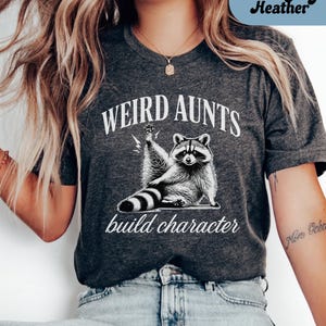 May include: Dark grey heather t-shirt with the text "WEIRD AUNTS build character" and a graphic of a raccoon raising its paw. The raccoon is black and white. The text is in white.