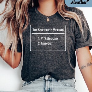 May include: Dark grey heather t-shirt with the text "The Scientific Method" and the steps "1. F**k Around 2. Find Out" in a white rectangular box. The shirt is worn by a person with long blonde hair and blue jeans.