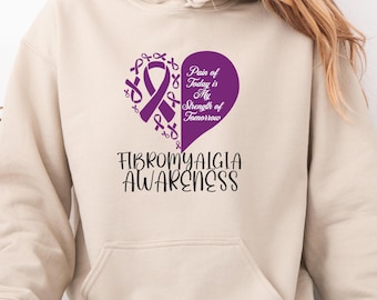 May include: A beige hoodie with a front pocket and drawstring hood. The front features a purple heart design with a ribbon and the text "Pain of Today is My Strength of Tomorrow." Below is the text "FIBROMYALGIA AWARENESS."