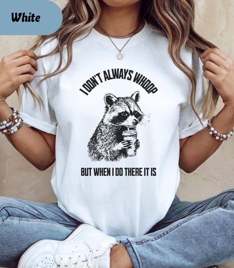May include: White t-shirt with a raccoon graphic holding a drink. The text reads "I DON'T ALWAYS WHOOP BUT WHEN I DO THERE IT IS" in black. The shirt is a casual style.