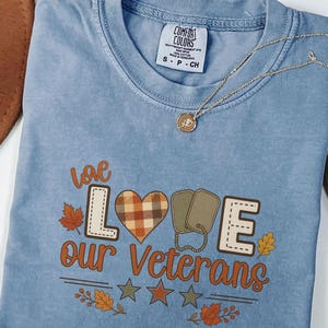 May include: A blue jean coloured t-shirt with a graphic print that reads "love our VETERANS". The design features a plaid heart, two olive green dog tags, autumn leaves, and three stars. A small gold necklace rests on the neckline of the shirt.