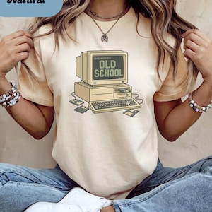 May include: A beige t-shirt with a retro computer graphic. The design features a monitor displaying "OLD SCHOOL", a keyboard, floppy disks, and a computer tower. The shirt has a crew neck and short sleeves.