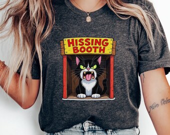 May include: Dark grey heather t-shirt featuring a cartoon cat in a wooden booth with the text "HISSING BOOTH". The cat is black and white with green eyes and its tongue sticking out. The shirt has a crew neck.