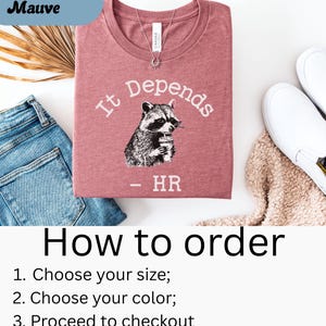May include: Heather mauve t-shirt with the text "It Depends" and a raccoon graphic holding a cup. The shirt also has the text "- HR" below the raccoon. The shirt is folded and placed on a white surface with blue jeans and white shoes.