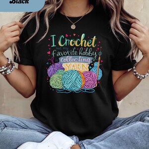 May include: Black t-shirt with the text "I Crochet but my favorite hobby is collecting YARN." The graphic features colorful yarn balls and a crochet hook. The shirt is a crew neck.