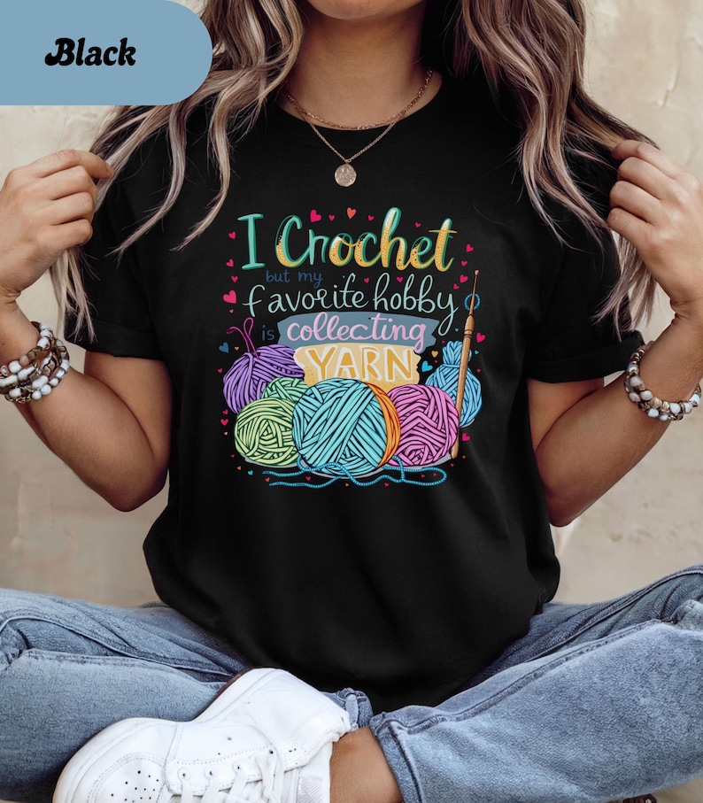 May include: Black t-shirt with the text "I Crochet but my favorite hobby is collecting YARN." The graphic features colorful yarn balls and a crochet hook. The shirt is a crew neck.