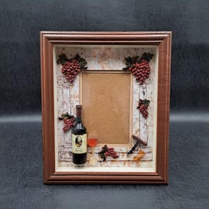 May include: A brown framed photo holder with a wine theme. The frame features a bottle of Shiraz wine, a wine glass, grapes, and a corkscrew. The background has a rustic, weathered look.