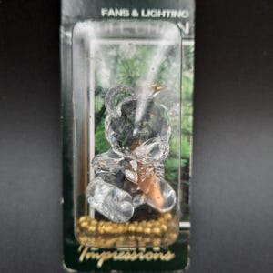 Könnte beinhalten: Eine klare Kristallbärenfigur in einer Kunststoff-Vitrine. Der Bär ist facettiert, mit einem goldfarbenen Sockel. Die Verpackung trägt die Aufschrift "FANS & LIGHTING" und "Impressions".