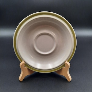 May include: A cream-colored saucer with a light green border. The saucer has a recessed center and is displayed on a wooden stand against a dark background. The design is simple and classic, suitable for everyday use.