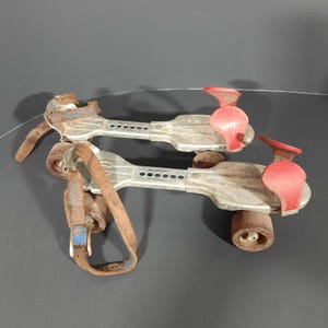 May include: A pair of vintage roller skates with metal frames and brown leather straps. The skates have red toe stops and rusty wheels. The metal frames show signs of wear and age, suggesting they are antique or retro.