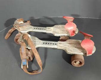 May include: A pair of vintage roller skates with metal frames and brown leather straps. The skates have red toe stops and rusty wheels. The metal frames show signs of wear and age, suggesting they are antique or retro.
