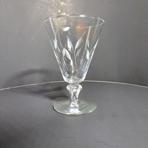 May include: A clear glass goblet with a delicate etched design of leaves. The goblet has a tall, slender stem and a round base.