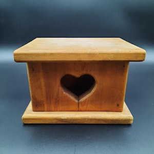 May include: A small, wooden box with a heart-shaped cutout on the front. The box is made of light-colored wood and has a rectangular shape with a flat top and base. The heart cutout adds a decorative touch to the rustic design.