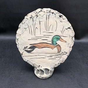 May include: A circular ceramic plaque featuring a relief carving of a mallard duck swimming in water with cattails in the background. The duck has a green head, brown wings, and a light-colored body. The plaque has a textured, rough-hewn border.