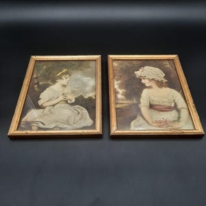 May include: Two framed antique prints featuring young women in period clothing. The prints are in gold-colored wooden frames. The artwork depicts the women in a soft, muted color palette, with a focus on their faces and attire.