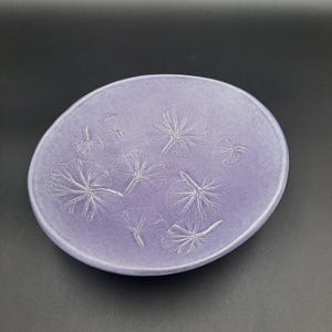 May include: A round, lavender-colored ceramic plate with embossed leaf designs. The plate has a slightly raised edge and a smooth surface. The leaf patterns are in a lighter shade, creating a subtle contrast. The plate is suitable for serving or display.
