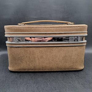 May include: A tan textured vanity case with a top handle and a horizontal silver-toned metal band across the front. The metal band features a central reflective panel and two keyhole locks, one marked with the letter M and the other with the letter H.
