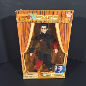Może przedstawiać: Kolekcjonerska marionetka Chrisa Kirkpatricka z *NSYNC, zapakowana w pudełko z motywem sceny. Lalka ubrana jest w czarno-czerwony strój. Pudełko zawiera logo *NSYNC i markę Living Toyz.