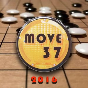 May include: A circular pinback button featuring a yellow textured background with the black text MOVE 37 and the red text 2016 below it. The button is positioned over a wooden board game surface with scattered black and white circular game pieces.