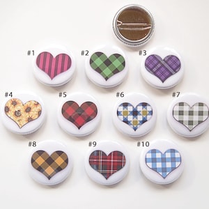 May include: A collection of ten heart-shaped button pins with various designs. The designs include stripes, plaid patterns in green, purple, red, and yellow, and a sunflower. The buttons are white with a metal pin backing.